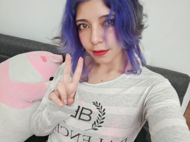 DarckVelvet with purple hair, wearing a gray striped shirt, making a peace sign, has red lipstick, sitting on a gray couch with a pink stuffed animal.
