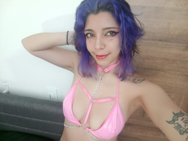 DarckVelvet with purple hair, pink strappy bikini top, showing tits, tattoos, nose ring, and chain, smiling at camera.