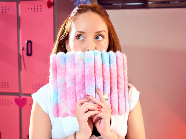 MollyMarmalade holding pink and blue fluffy pasties over her tits, with red nails, in a locker room. She has brown hair and blue eyes.
