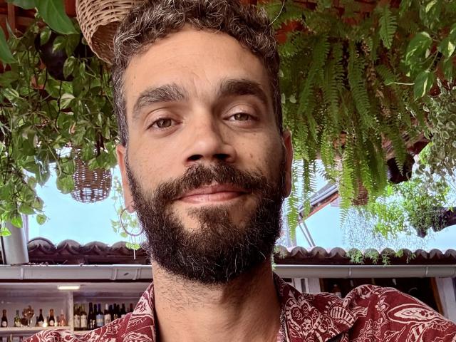 HairyBreeder has curly brown hair, a thick black beard, and is smiling slightly. He's wearing a red patterned shirt. Green plants hang above him.