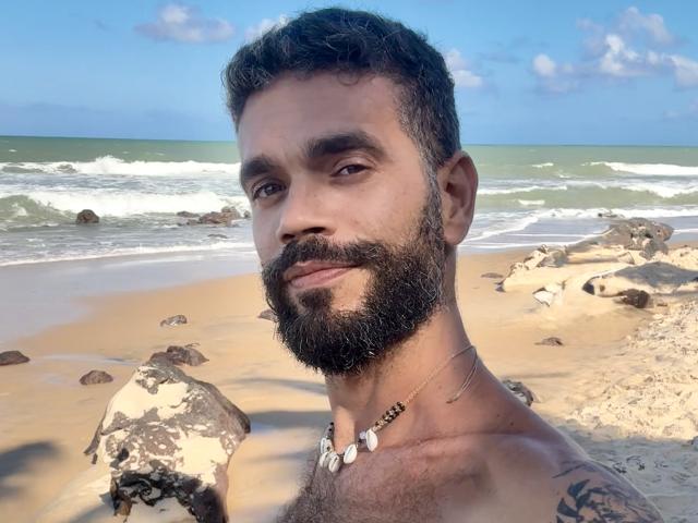 HairyBreeder has a dark, curly beard, chest hair, and a necklace with white beads. He's shirtless with tattoos, standing on a sandy beach with rocks and ocean.