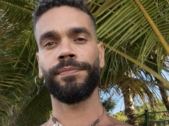 HairyBreeder has a dark, thick beard, brown eyes, and short curly black hair. He's wearing a silver hoop earring and a chain necklace, smiling slightly.
