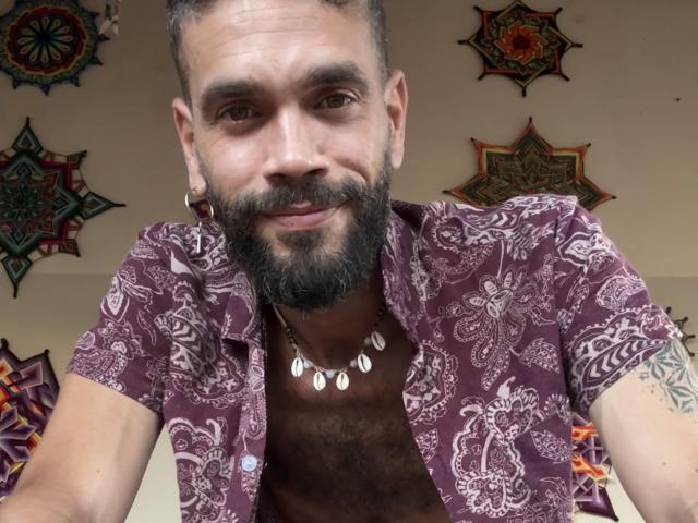 HairyBreeder with a beard, wearing a purple patterned shirt, necklace with shell charms, and hoop earrings, smiling at the camera. His chest is hairy and he has tattoos.
