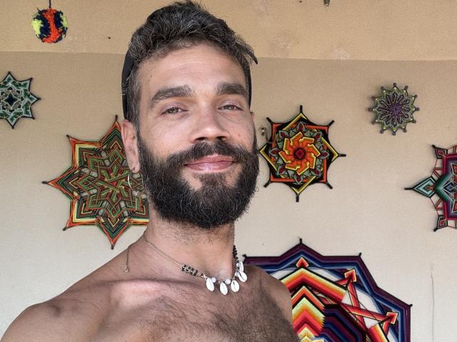HairyBreeder, shirtless with a full beard, necklace, and dark hair, smiling at the camera. Boobless, hairy chest, and a relaxed, confident vibe. Decorative, colorful wall art in the background.