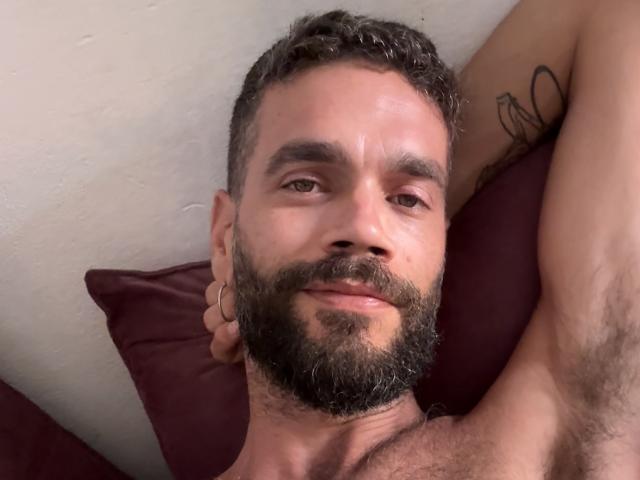 HairyBreeder has a beard, short curly hair, and a tattoo on his arm. He's lying down with a ring on his finger, smiling with his arm behind his head.