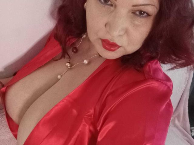 MelissaRilley with red lipstick, wavy dark hair, wearing a red satin robe, showing her big tits, necklace with pearls, and a slightly bored expression.