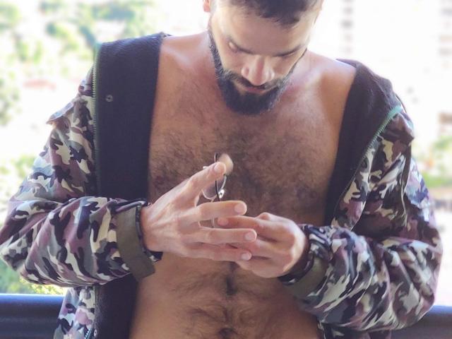 HairyBreeder, shirtless with a hairy chest, holding a small, erect, circumcised penis in his hands, wearing a camouflage jacket with black straps, looking down.