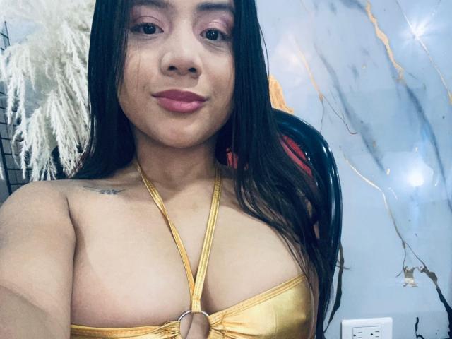 MaddieEvans with long black hair, wearing a gold halter top that exposes her tits, has pink lipstick, and a small tattoo on her left shoulder. She's smiling.
