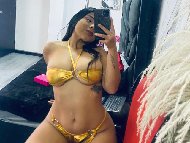 MaddieEvans in a shiny gold bikini, showing her tits with an O-ring, holding a phone, sitting on a bed, long black hair, tattoo on right arm, mirror selfie.