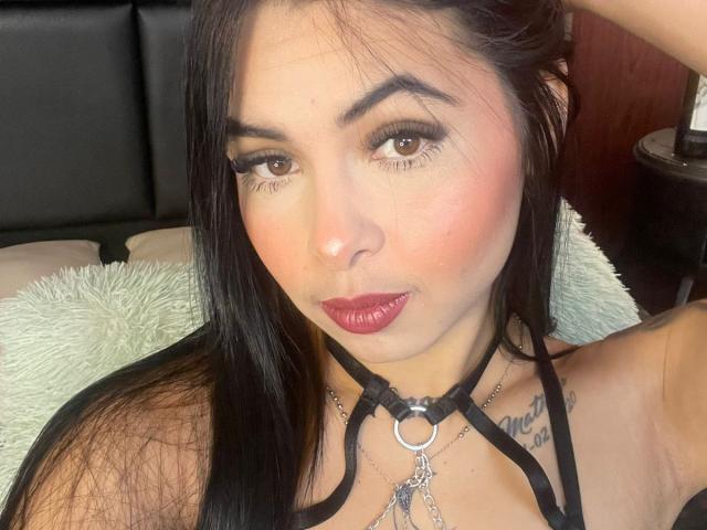 MistressAsshley has black hair, brown eyes, and dark red lipstick. She's wearing a black halter top with a metal ring and chain collar, showing her tits and tattoo.