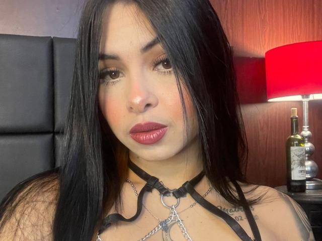 MistressAsshley has long black hair, red lipstick, and wears a black leather choker with metal rings, looking at the camera with a slight smirk. Her tits are visible.