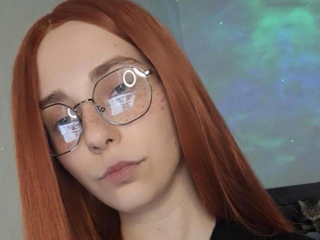 EvaLoraveiI has red hair, clear glasses, and freckles. She's looking slightly to the side with a neutral expression, and her lips are slightly parted.