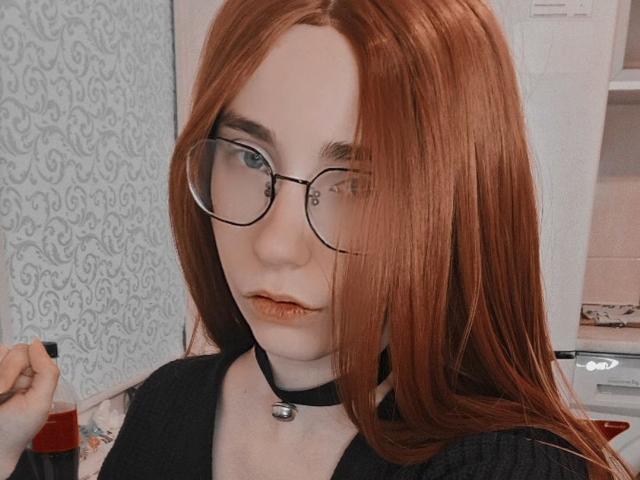 EvaLoraveiI with red hair, glasses, black choker, and black top, holding a red bottle, has pale skin and pouty lips, looking directly at the camera.