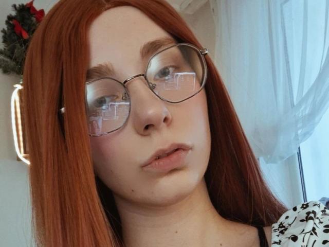 EvaLoraveiI has red hair, glasses, and pouty lips, looking serious. Her tits are small, and her pussy is not visible. She's wearing a black and white patterned top.