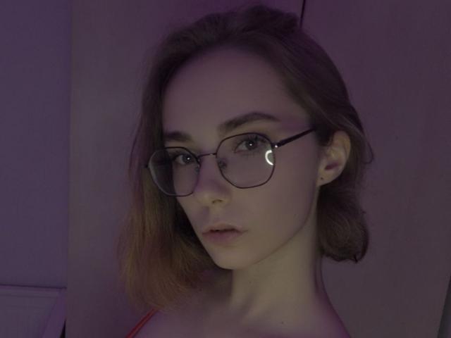 EvaLoraveiI has light skin, brown shoulder-length hair, and wears round glasses with a crescent moon on the right lens. She's looking at the camera with a neutral expression.