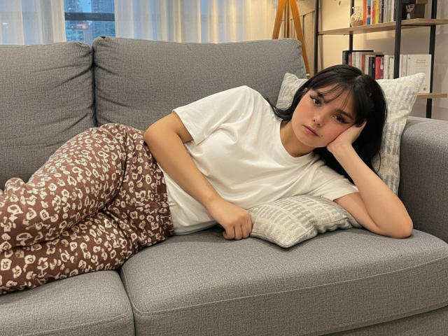 AbbyStanley, with black hair and bangs, lies on a grey couch in a white t-shirt and brown patterned pajama pants, resting her head on a pillow.