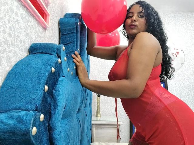 LiliLopez with curly black hair, wearing a red dress, leaning on a blue, tufted, stuffed chair, holding red balloons, showing her ass and tits, in a brightly lit room.