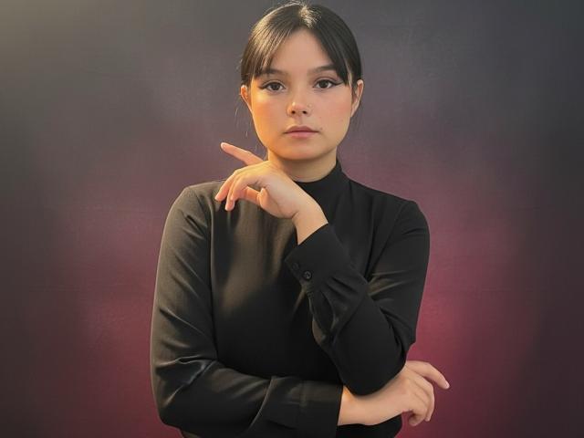 AbbyStanley with dark hair, wearing a black long-sleeve top, resting chin on her hand, looking serious, arms crossed, soft lighting, subtle pink and purple background.