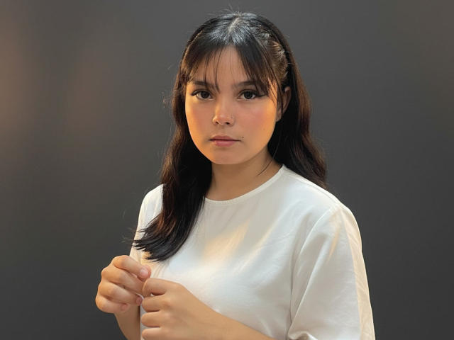 AbbyStanley with black hair, bangs, and a white shirt, holding her shirt with both hands, looking slightly annoyed, medium tits, dark background, soft lighting, serious expression.