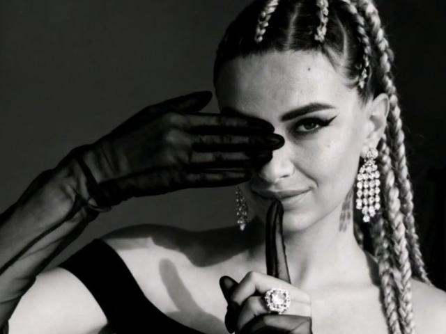 Black and white portrait of PassionX. She's got braided hair, thick and straight, falling to the sides. She's got dark, dramatic eyeliner and is looking to the side with a slight smirk. She's wearing a black off-the-shoulder top, and sheer black gloves. Her right hand is up, covering her eye, while her left hand holds a black ribbon. She's got dangly earrings and a ring with a big stone on her left hand. The texture of her hair and the sheer gloves are highlighted. The composition is focused on her face and upper chest.