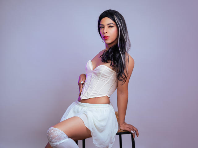 KhendraGrey with black hair, white corset, and skirt, sitting on stool, showing white thigh-high stockings, and a subtle sideboob, with a confident, sexy expression.
