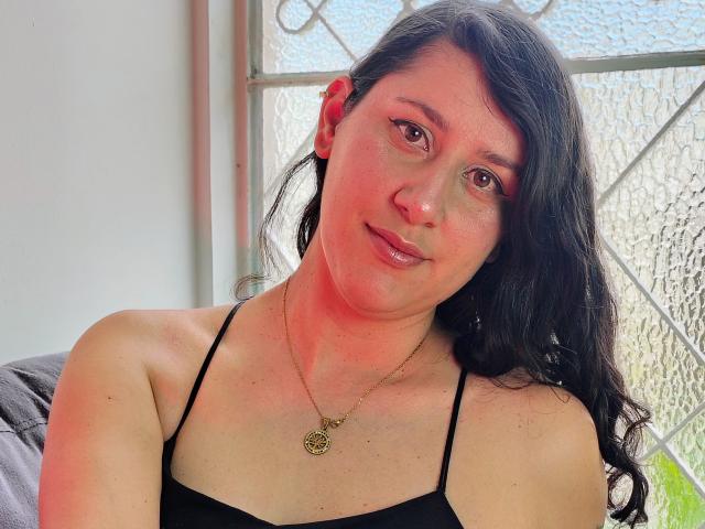 LunaDamasco has red-tinged skin, long black hair, brown eyes, and wears a black spaghetti-strap top with a gold necklace, smiling softly at the camera.