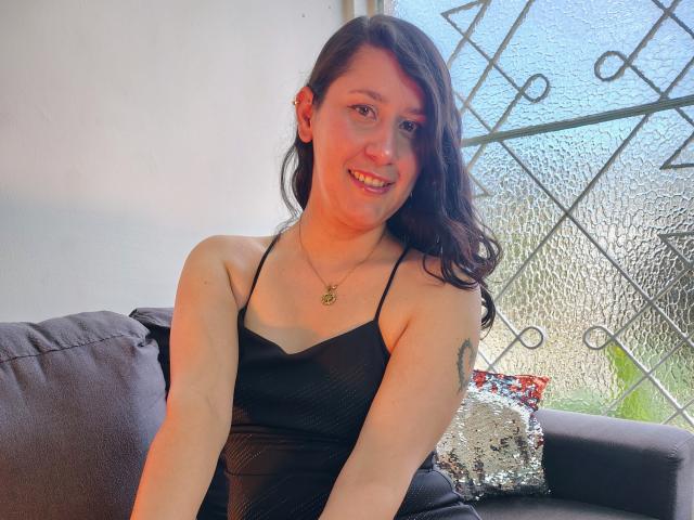 LunaDamasco, a light-skinned woman with long brown hair, sits on a dark gray couch. She wears a black, spaghetti-strap, textured dress and a gold necklace with a small pendant. She has a tattoo on her right arm and smiles at the camera. A silver, sequined pillow is on the couch's right side. The background includes a textured glass window with a metal grid pattern. Bright natural light is coming from the window.