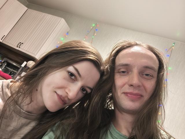KissySmell with long brown hair and KissySmell with long brown hair and a slight smile, both in a kitchen with colorful string lights in the background.