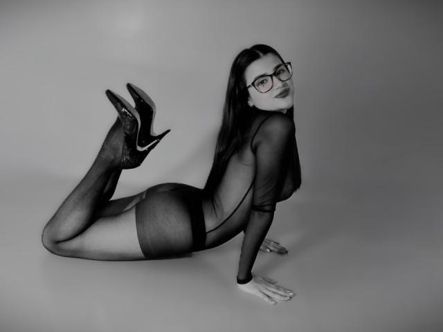 IrisBlair in a sheer black dress, on all fours, with her ass up, wearing black high heels and glasses, looking back over her shoulder.