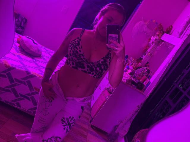 Mirror selfie of SaritaCaramelo with purple lighting. She has light skin, blonde hair, wearing a leopard-print bra, and white pants with floral patterns. She holds a smartphone in her right hand, covering part of her face. The room has a bed with a geometric patterned cover and a cluttered shelf with various items in the background. Her midriff is exposed, showing a slight belly. The image is casual and focused on her upper body.