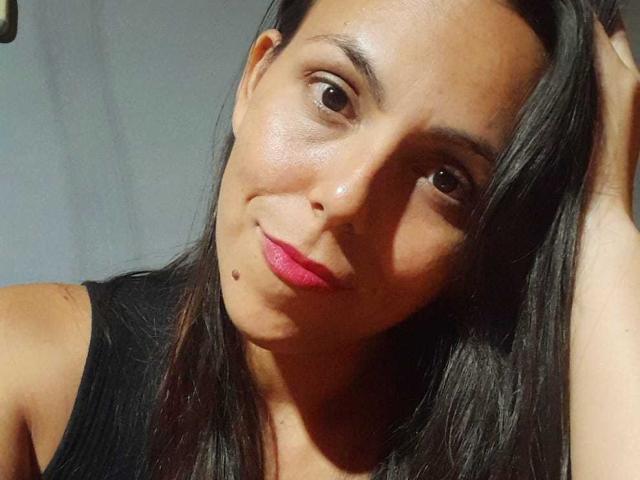 AfroditSexx with medium brown skin, black straight hair, pink lipstick, small black mole on chin, brown eyes, wearing black tank top, slightly smiling, head tilted right.