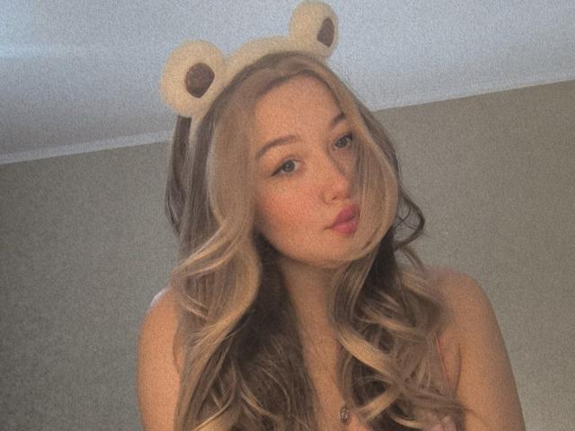 CloraFlippin with long blonde and brown wavy hair, wearing a cute bear-eared headband, pouting lips, and no top, in a soft-lit, slightly grainy photo.