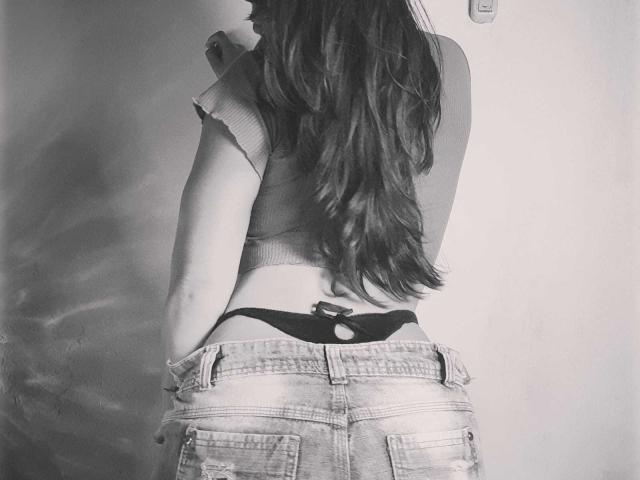 Black-and-white photo of AfroditSexx from behind. She has long, wavy hair flowing down her back. She's wearing a cropped, short-sleeve top and distressed jeans. Her black thong is visible, partially tucked into her jeans. The wall in the background is plain, with a small, square light switch to the right. The texture of her hair contrasts with the rough, worn denim. The image focuses on her lower back and buttocks.
