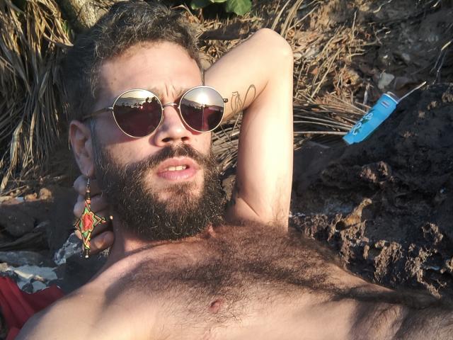 HairyBreeder with a beard, wearing reflective sunglasses, lying on a towel, exposing hairy chest, holding a colorful earring, and looking annoyed. Blue object to the right.