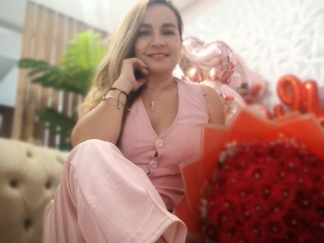 ManuelaMature with blonde hair, pink button-up dress, and bracelet, smiling, holding pink balloons, sitting on beige couch, leg up, red rose bouquet in foreground.