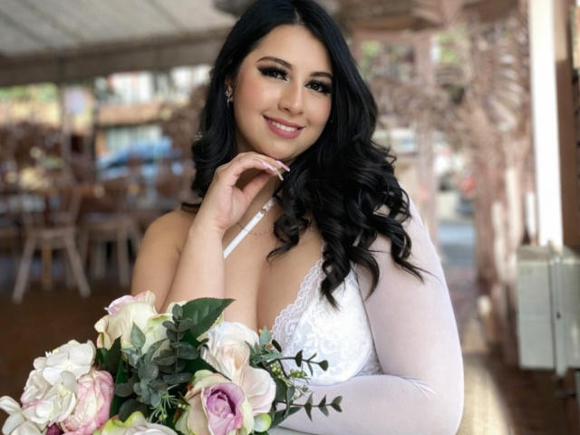 AshleeyPrinces with long black hair, white lace dress, and big tits, holding a bouquet of pink and white roses, smiling at the camera.