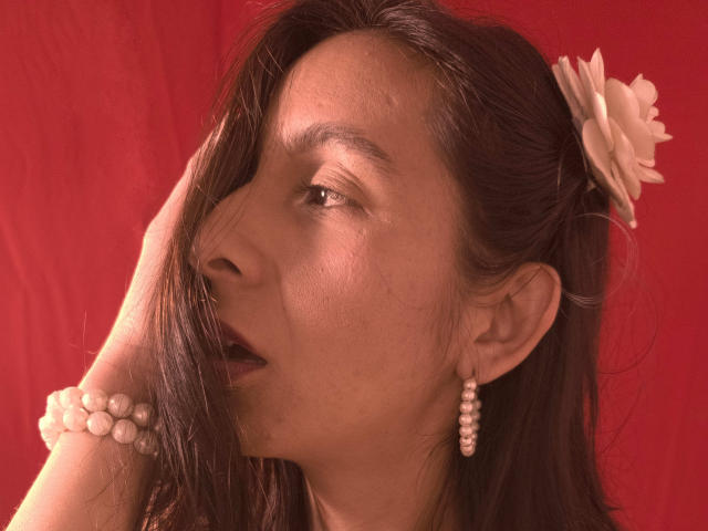MiaAmoree has long brown hair with a white flower, wearing pearl earrings and bracelet, hand in hair, red background, looking to the side with slightly open mouth.