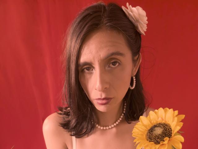 MiaAmoree with shoulder-length brown hair, white flower in hair, pearl necklace, hoop earrings, and sunflower in hand, looking serious against red background. Boobs visible, no makeup.