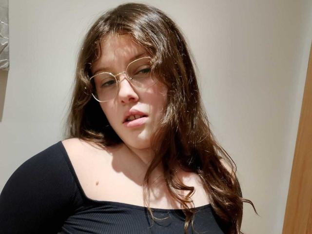 XSari has wavy brown hair, glasses, and a black off-shoulder top, looking slightly open-mouthed with a small mole on her neck. Her tits are small and she's fair-skinned.