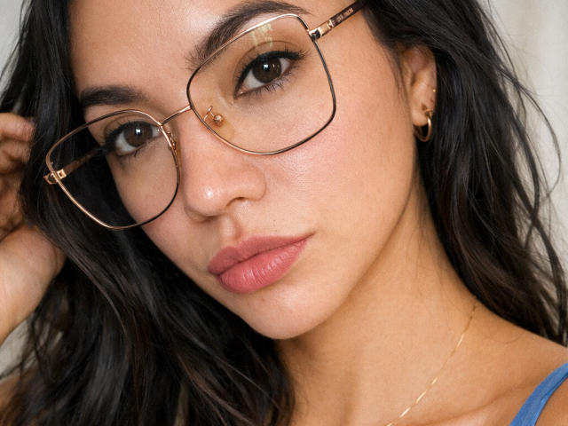 SofieWhite has brown eyes, wears big gold-rimmed glasses, and has full pink lips. She's a cute brunette with slightly wavy hair, a gold hoop earring, and a blue top.