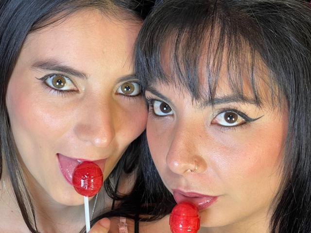 Close-up photo of ValkiriaAndDione, two women with dark hair and striking eye makeup. Both have fair skin and are holding red lollipops in their mouths. ValkiriaAndDione on the left has her tongue slightly out, while the one on the right has a more subtle, closed-mouth expression. Both have black eyeliner and are looking directly at the camera. ValkiriaAndDione on the right has a small nose ring. The image has a sharp focus on their faces, with a soft, natural light highlighting their skin and hair. The lollipops are shiny and red, contrasting with their pale skin.