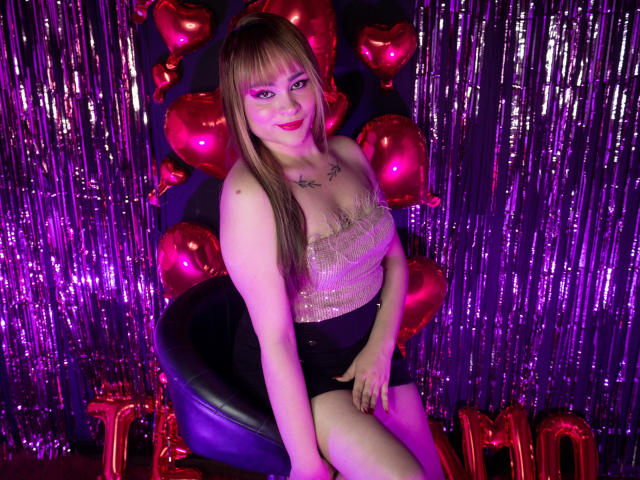LailaDurmont with straight brown hair, pink makeup, sparkly top, black shorts, sitting on purple chair, surrounded by red heart balloons and purple foil, smiling at camera.