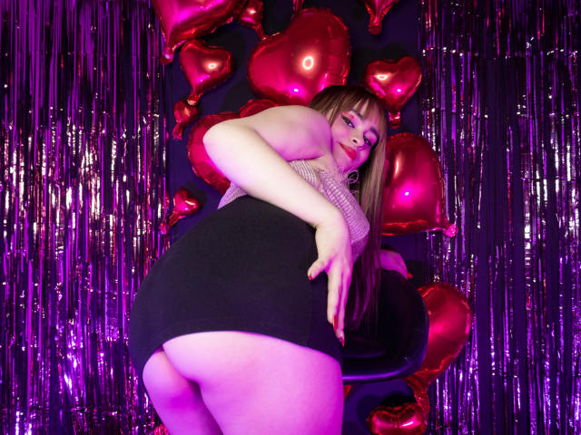 LailaDurmont with big ass in a black skirt, her butt exposed, wearing a fishnet top, standing in front of pink heart balloons and purple string, looking back.
