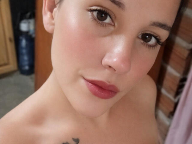Maruli's face with bare shoulders, light skin, brown eyes, pink lipstick, small tattoo on chest, short brown hair, and subtle makeup, looking slightly upward.