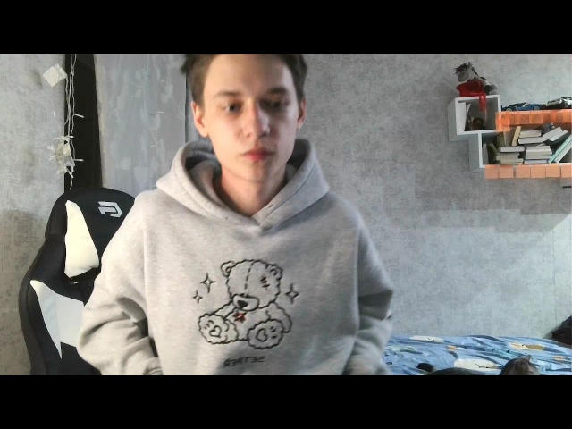 Gornits with short brown hair, wearing a gray hoodie with a cute bear and wheels design, sitting in a chair, looking slightly down, in a dimly lit room.