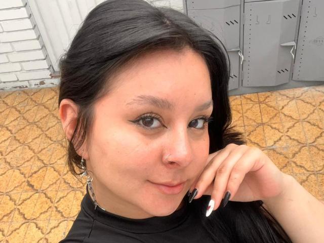 EmaJames tiene el pelo negro, un piercing en la nariz y delineador de ojos negro. Está tocándose la barbilla con esmalte de uñas negro y blanco, y lleva una camiseta negra. Sus senos no son visibles..