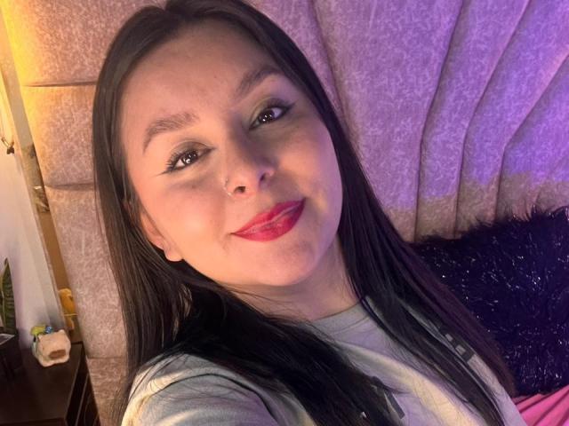 EmaJames with long black hair, red lipstick, and eyeliner, smiling at the camera, wearing a gray shirt, nose ring, and light skin. Her tits are not visible.