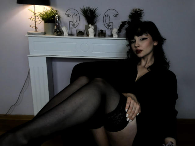 DeniseKitty, con i capelli neri, il rossetto scuro e la lingerie nera, seduta con le gambe incrociate, che mostra la sua biancheria intima in pizzo nero e le calze nere trasparenti, con le braccia appoggiate sulle cosce..