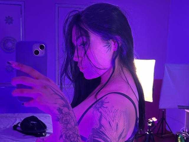 KatianPierce with long black hair, purple-lit, tattooed arm, wearing a black spaghetti strap, taking a selfie in a mirror. Boobs visible, side profile, holding a purple phone.