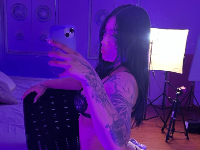 KatianPierce with long black hair, glasses, and tattoos, taking a selfie in purple lighting, wearing black lace bra, kneeling on bed, holding black chair with white dots.